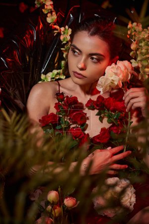attractive young woman in wet red dress lying in black bathtub among blooming flowers
