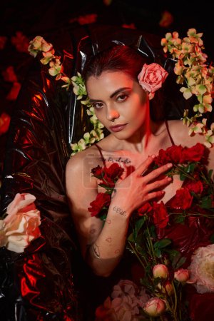 tattooed woman in wet red dress looking at camera and lying in bathtub among blooming flowers