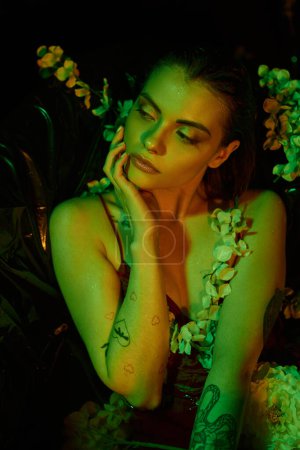grüne Licht, attraktive junge Frau mit nassen Haaren berührt Gesicht und posiert zwischen Blumen