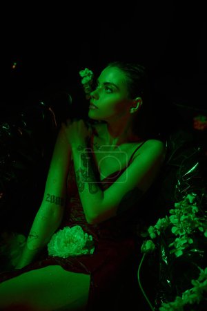 grüne Licht, sinnliche und elegante Frau mit nassen Haaren, die wegschaut und zwischen Blumen posiert