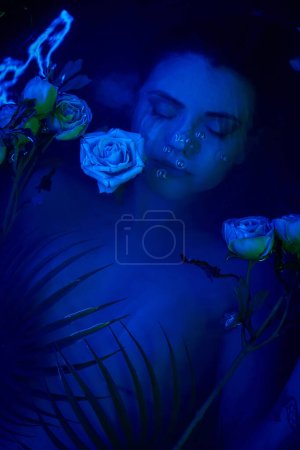 belleza femenina, mujer joven buceando bajo el agua entre hojas de palma y flores, luz azul