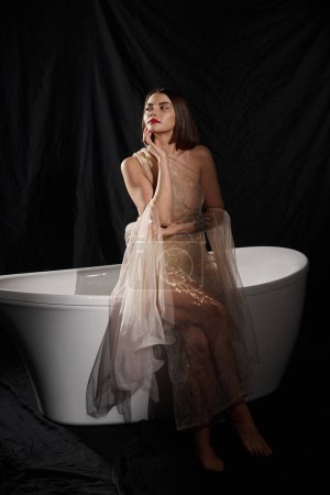 feminine elegance, barefoot young woman in romantic dress standing near white bathtub on black