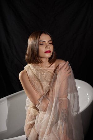 feminine sensuality, dreamy young woman in transparent dress standing near white bathtub on black