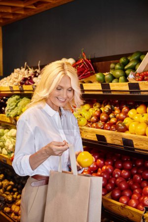 lustige reife Frau in Freizeitkleidung, die im Supermarkt gelbe Tomaten in die Einkaufstasche steckt