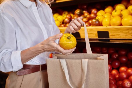 vista recortada de la mujer madura poniendo tomate amarillo en la bolsa de compras, mientras que en la tienda de comestibles