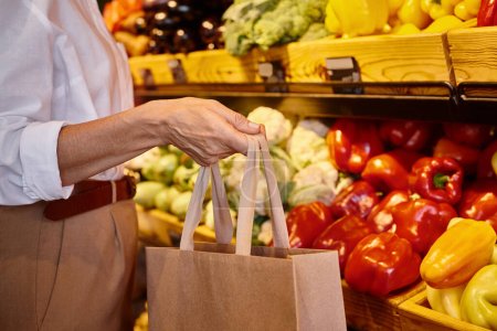 cropped view of mature stylish woman in casual attire holding shopping bag at grocery store