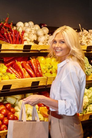 vertical shot of mature woman with shopping back smiling at camera with vegetable stall on backdrop