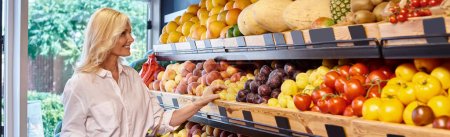 joyful mature woman with blonde hair in casual outfit choosing fruits at grocery store, banner