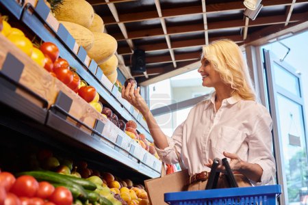 mujer madura feliz en traje casual con cesta de la compra en las manos la elección de las frutas en la tienda de comestibles