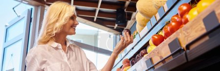 mature woman in casual outfit with shopping basket in hands choosing fruits at grocery store, banner