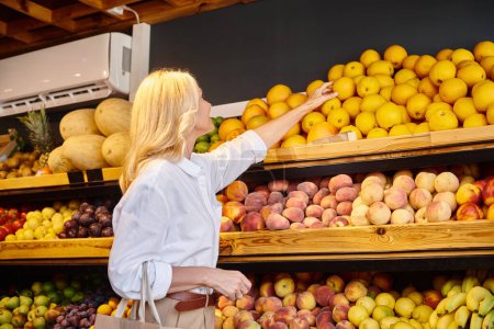 Foto de Madura rubia alegre mujer en la ropa cotidiana con bolsa de compras recoger naranjas en la tienda de comestibles - Imagen libre de derechos