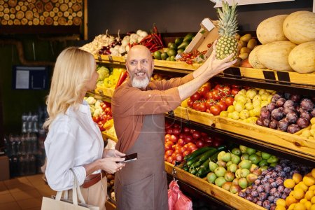 Téléchargez les photos : Vendeur barbu joyeux montrant l'ananas énorme à son client féminin à côté de l'épicerie étal - en image libre de droit