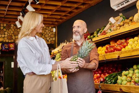 cheerful bearded seller giving fresh pineapple to his mature jolly female customer at grocery store