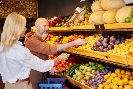 focus on bearded mature seller helping his blurred senior female customer to choose fruits