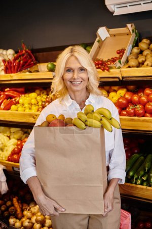 Foto de Mujer madura de buen aspecto en traje casual sosteniendo bolsa de compras llena de frutas y sonriendo felizmente - Imagen libre de derechos