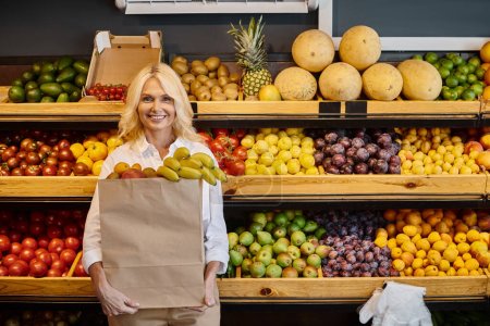 Foto de Alegre atractiva mujer madura en ropa cotidiana sonriendo y sosteniendo bolsa de compras con plátanos - Imagen libre de derechos