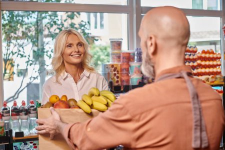 focus on cheerful mature female customer smiling at blurred seller with shopping bag full of fruits