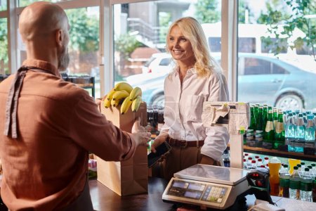 focus on jolly mature customer smiling at blurred seller next to cash desk at grocery store