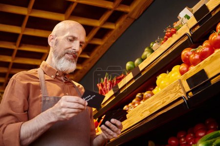 mature seller with beard holding price tags posing with grocery stall with vegetables on backdrop