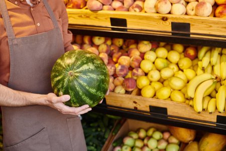 cropped view of mature seller holding fresh juicy watermelon with grocery stall on background