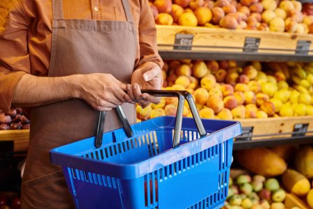 cropped view of mature salesman holding shopping basket in hands with blurred fruits on background