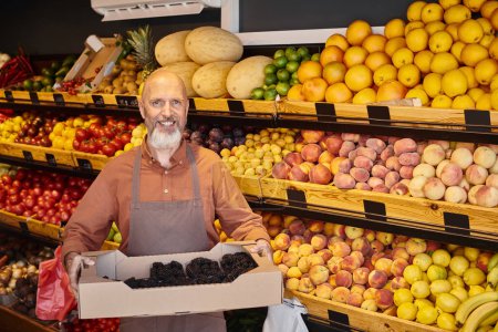 Foto de Alegre gris barbudo vendedor posando con frescas y deliciosas moras en las manos y sonriendo alegremente - Imagen libre de derechos