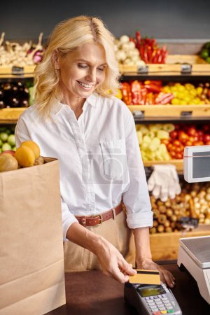 mature happy female customer with shopping bag with fruits on cash desk paying with her credit card