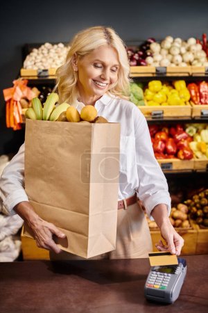 Foto de Alegre mujer madura en traje casual sosteniendo bolsa de compras y pagando con su tarjeta de crédito - Imagen libre de derechos