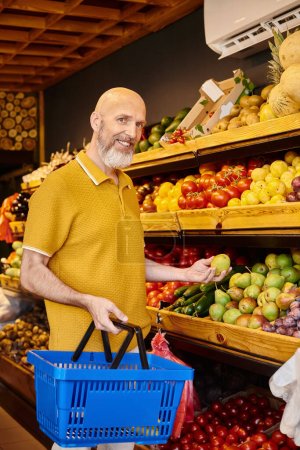 Foto de Alegre gris barbudo cliente masculino recogiendo frutas frescas y sonriendo a la cámara en la tienda de comestibles - Imagen libre de derechos