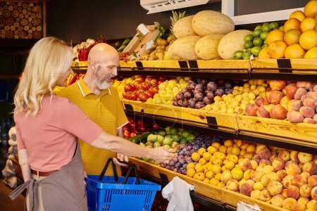 Foto de Alegre vendedora madura ayudando a su cliente barbudo a elegir frutas nutritivas en la tienda de comestibles - Imagen libre de derechos