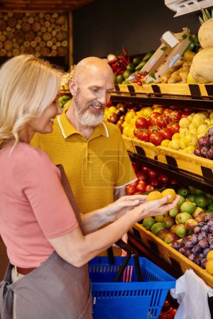 focus on joyous mature man with shopping basket getting help from saleswoman while choosing fruits
