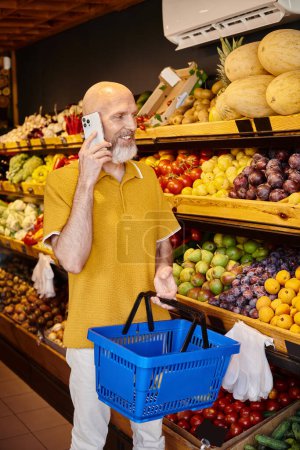 Foto de Buen aspecto cliente varón maduro con cesta de la compra hablando por teléfono al elegir comestibles - Imagen libre de derechos