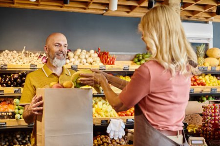 Photo for Focus on jolly customer smiling next to blurred female seller putting fresh bananas into paper bag - Royalty Free Image