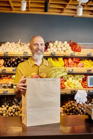 cheerful bearded man posing with paper bag full of fresh fruits and smiling at camera at grocery