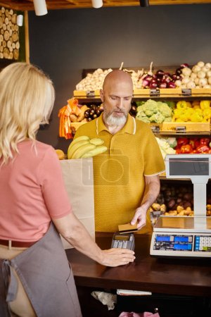 Foto de Cliente barbudo concentrado que paga con tarjeta de crédito por frutas al lado del vendedor maduro en el supermercado - Imagen libre de derechos