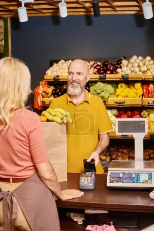 focus on jolly bearded man paying with his phone for fresh groceries and smiling at saleswoman