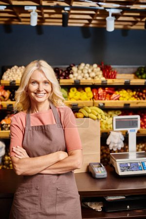 joyous blonde mature saleswoman posing with arms crossed on chest and smiling happily at camera