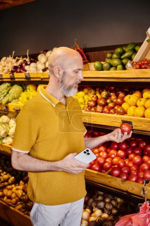 good looking mature man with beard holding mobile phone and looking at fresh tomato at grocery store