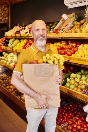 mature cheerful customer holding paper bag full of delicious juicy fruits and smiling at camera