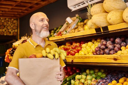 Foto de Cliente masculino maduro alegre posando con la bolsa de papel llena de frutas vibrantes frescas y mirando hacia otro lado - Imagen libre de derechos