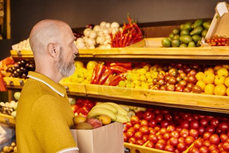 Foto de Hombre barbudo gris maduro concentrado sosteniendo bolsa de papel llena de frutas frescas en la tienda de comestibles - Imagen libre de derechos