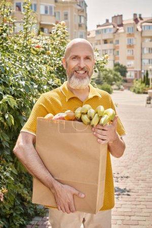 gai mature mâle client posant à l'extérieur avec sac en papier plein de fruits et souriant à la caméra