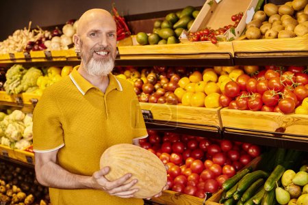 Téléchargez les photos : Homme barbu gris joyeux en tenue décontractée posant avec un énorme melon dans les mains et souriant à la caméra - en image libre de droit