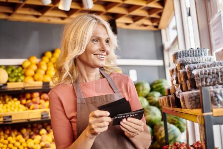 jolly mature seller holding price tags in hands smiling happily and looking away at grocery store