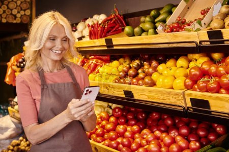 Foto de Alegre madura vendedora sonriendo felizmente y mirando su teléfono móvil en la tienda de comestibles - Imagen libre de derechos