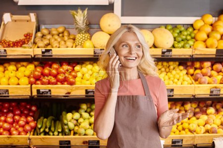 Foto de Alegre vendedor atractivo sonriendo y hablando por teléfono con frutas y verduras en el telón de fondo - Imagen libre de derechos