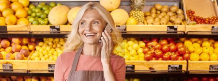 Foto de Feliz vendedor femenino sonriendo y hablando por teléfono con frutas y verduras en el telón de fondo, pancarta - Imagen libre de derechos