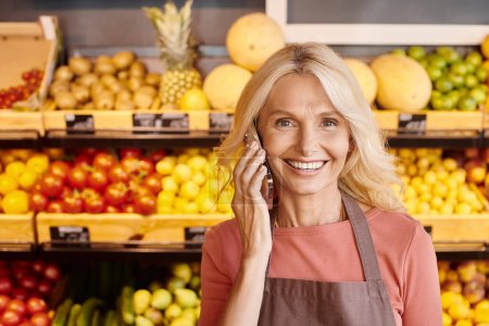 fröhliche reife blonde Verkäuferin, die mit dem Handy spricht und im Supermarkt in die Kamera lächelt