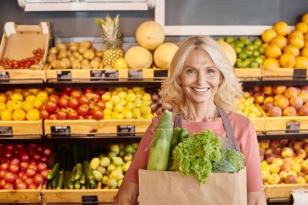 Foto de Alegre vendedor maduro con bolsa de papel llena de verduras en las manos sonriendo a la cámara en la tienda de comestibles - Imagen libre de derechos