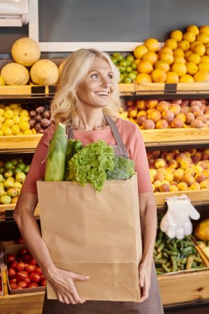Foto de Madura vendedora alegre posando con bolsa de papel llena de verduras y mirando hacia otro lado en la tienda de comestibles - Imagen libre de derechos